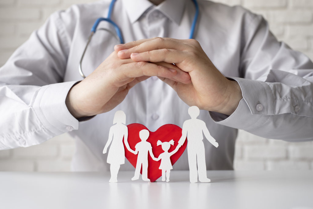 Um médico segurando as proprias mãos, mostrando uma familia de papel na frente de um coração de papel