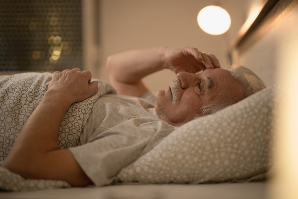 Homem idoso preocupado e cansado deitado na cama, mostrando que a Falta de sono pode envelhecer