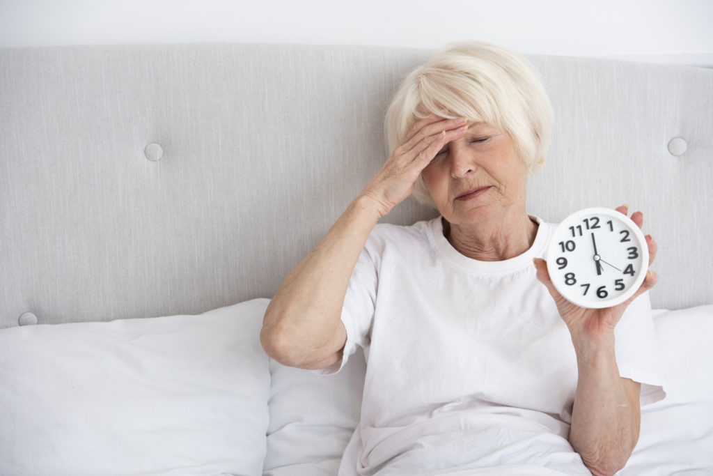 Senhora idosa usando uma camisa branca de manga, sentada na cama com uma expressão de cansaço, colocando a mão direita no rosto e usando a outra mão para segurar um relógio mostrando que a Falta de sono pode envelhecer