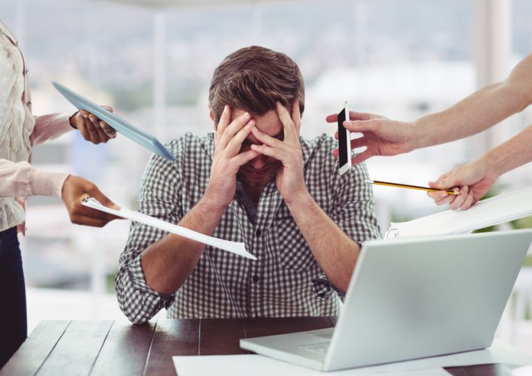 Homem estressado e frustrado com as duas mãos no rosto, na frente do notebook, enquanto muitas pessoas tentam entregar blocos, celulares e objetos de trabalho, estresse