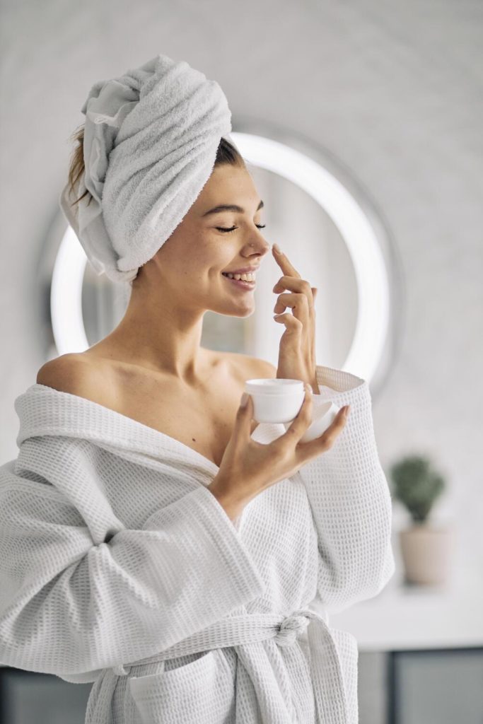 jovem branca e morena fazendo sua rotina matinal passando creme no rosto, ela veste um roupão e usa toalha no cabelo , autocuidado