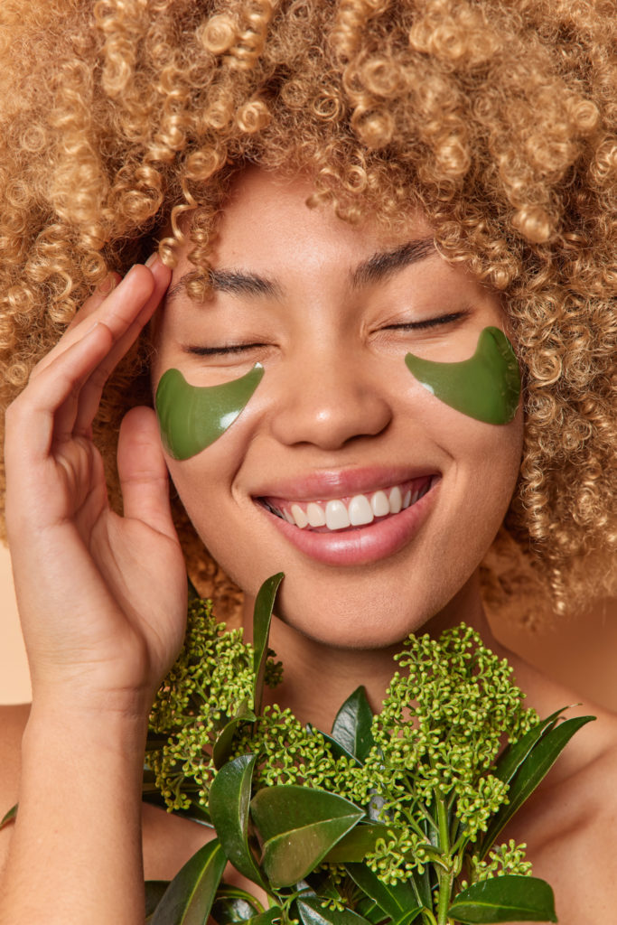 close, retrato de sorrisos de mulher de cabelos cacheados amplamente mostra dentes brancos aplica manchas verdes para reduzir o inchaco sofre procedimentos de beleza mantem os olhos fechados cuidados com a pele, beleza sustentavel